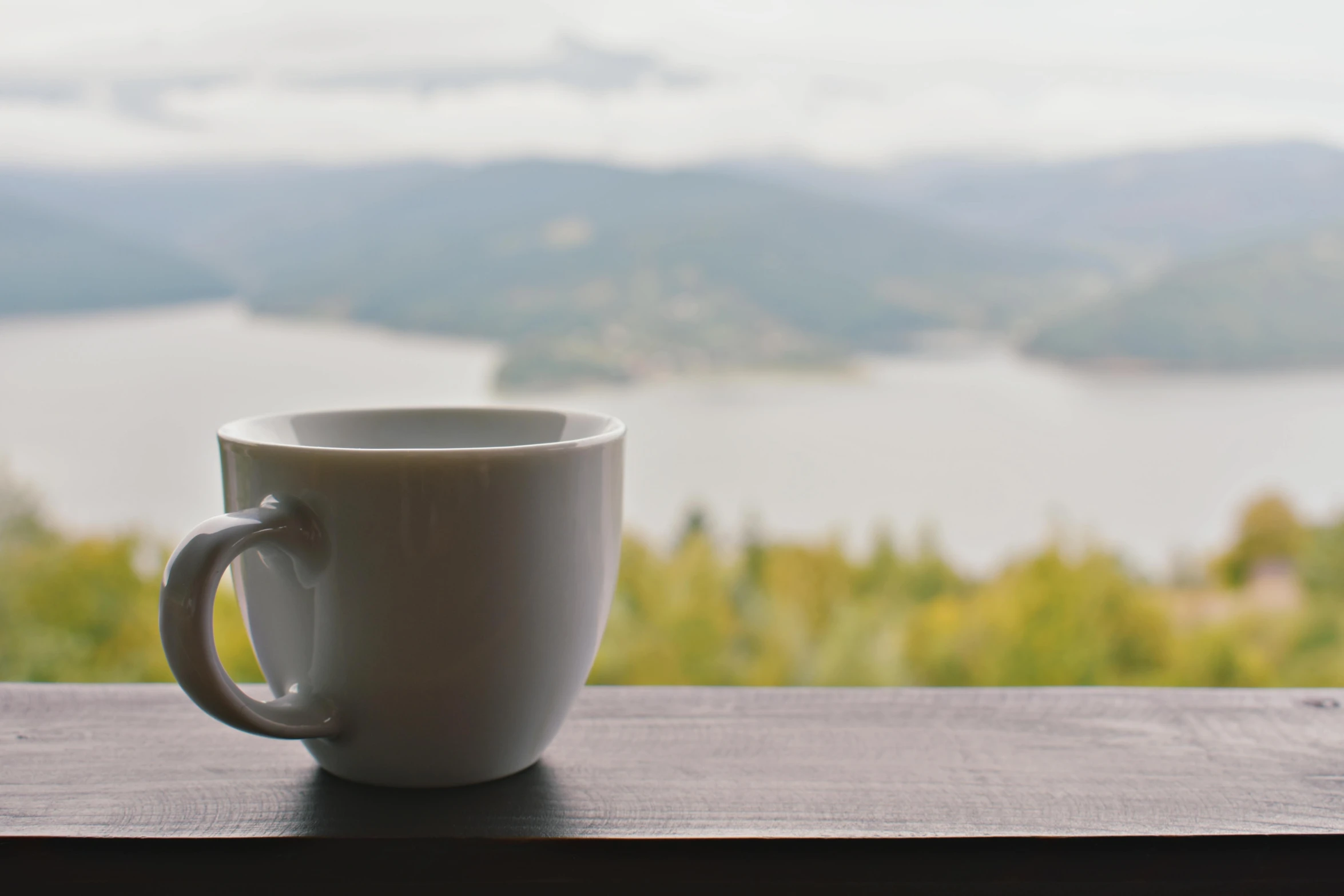 a white coffee cup sitting on top of a wooden table, inspired by Muggur, pexels contest winner, it has a lake in the distance, overlooking a valley, grey, whistler