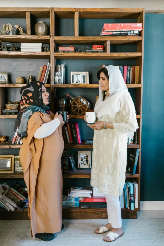 two women standing next to each other in front of a bookshelf, by Riza Abbasi, hurufiyya, wearing white robe, in a living room, wedding, inspect in inventory image