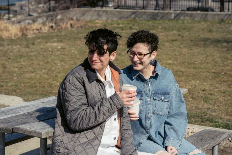 a couple of women sitting on top of a wooden bench, next to a cup, non-binary, humans of new york style, warm spring