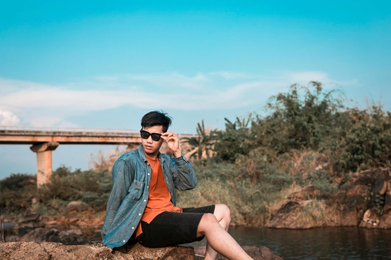 a man sitting on a rock next to a body of water, a picture, unsplash, realism, wearing orange sunglasses, avatar image, asian male, male teenager
