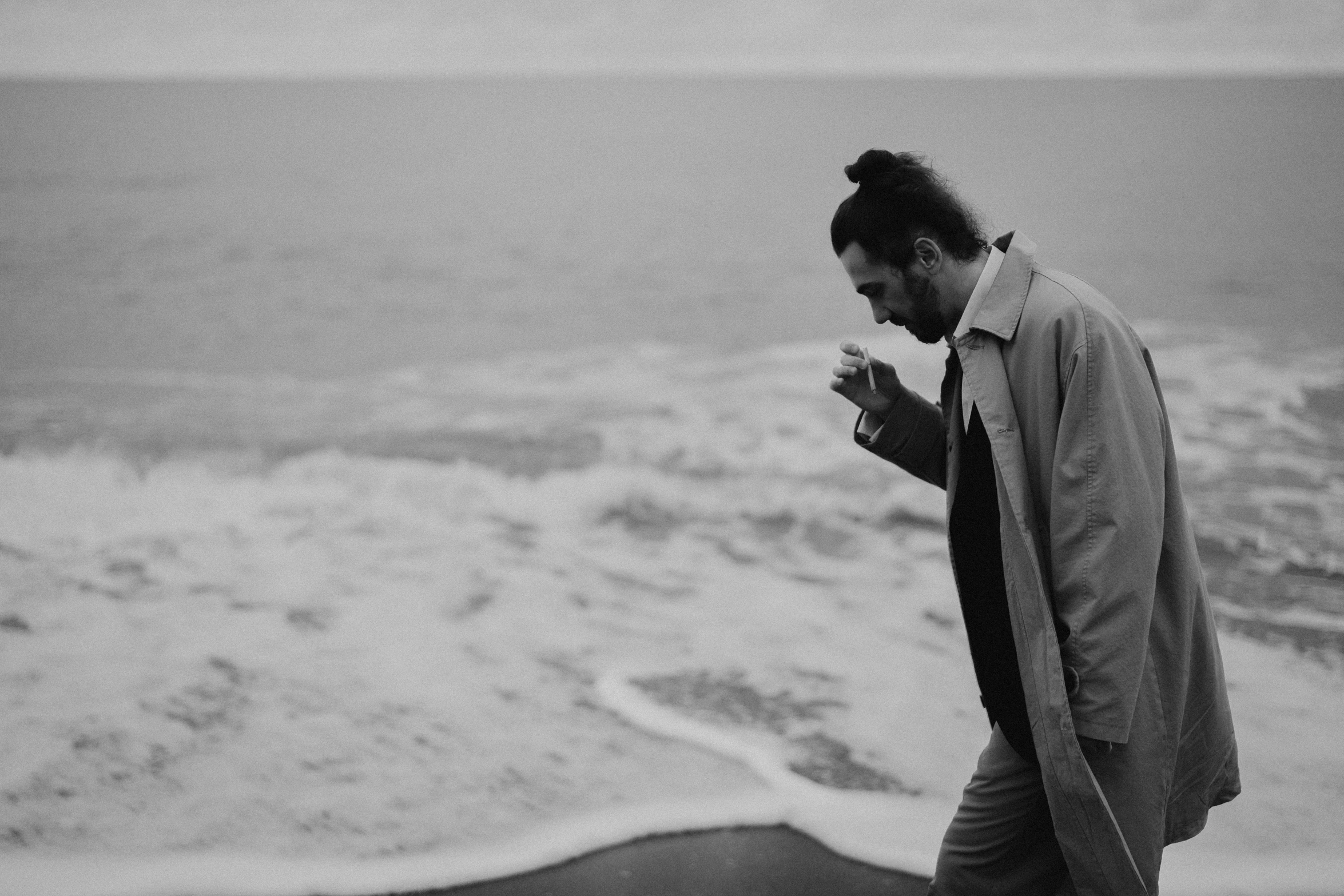 a man standing on top of a beach next to the ocean, a black and white photo, by Romain brook, unsplash, he is wearing a trenchcoat, holding a drink, swirling around, orelsan