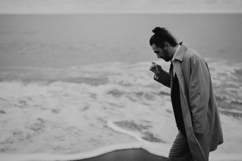 a man standing on top of a beach next to the ocean, a black and white photo, by Romain brook, unsplash, he is wearing a trenchcoat, holding a drink, swirling around, orelsan