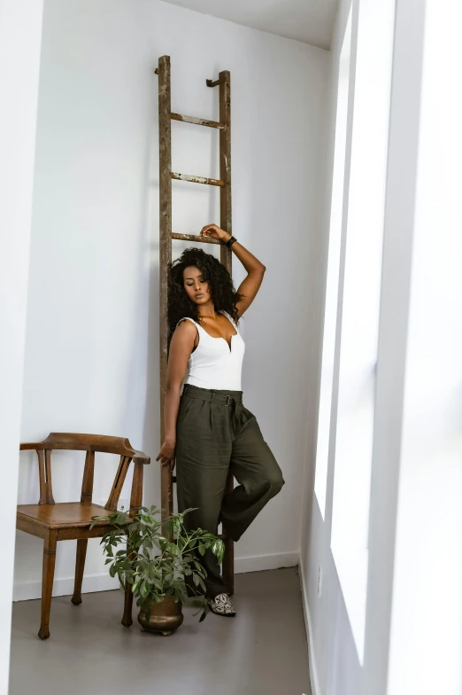 a woman standing next to a ladder in a room, inspired by Esaias Boursse, trending on unsplash, olive green slacks, in front of white back drop, wearing a camisole, dark-skinned