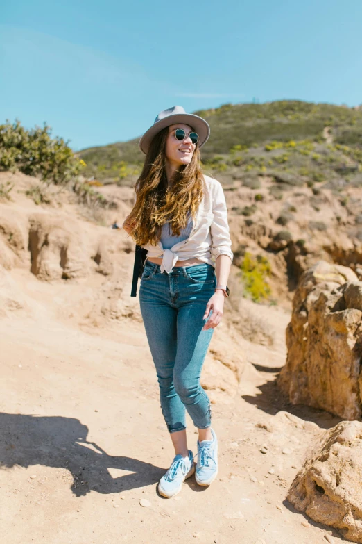 a woman standing on top of a rocky hill, by Gwen Barnard, trending on unsplash, tight blue jeans and cool shoes, wearing sunglasses and a hat, long flowy hair, blue fedora