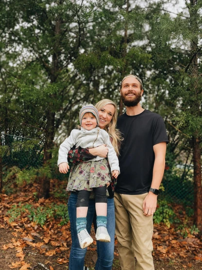 a man standing next to a woman holding a baby, by Winona Nelson, unsplash, against the backdrop of trees, bearded and built, husband wife and son, taken on iphone 14 pro