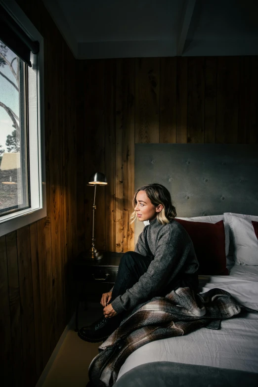 a woman sitting on a bed looking out a window, a portrait, by Lee Loughridge, unsplash contest winner, cabin lights, “ iron bark, hotel room, winter setting