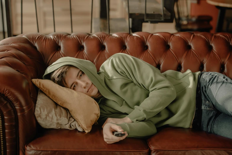 a woman laying on a couch with a remote in her hand, inspired by Elsa Bleda, trending on pexels, happening, green hood, sad man, beige hoodie, julian ope