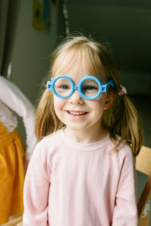 a little girl sitting on top of a wooden chair, wearing thin large round glasses, in blue and yellow clothes, zoomed in shots, kids playing