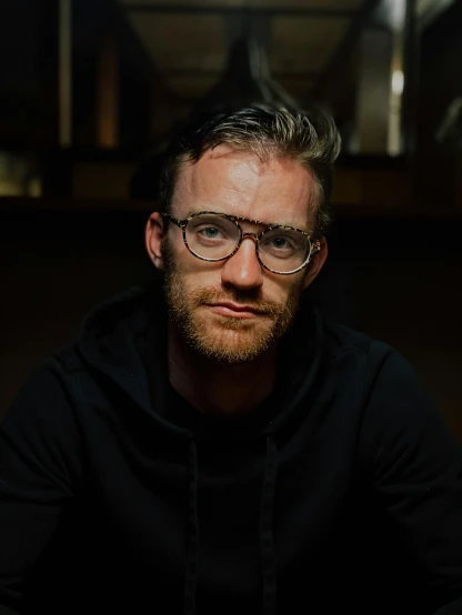 a man with glasses sitting at a table, a character portrait, by Jacob Toorenvliet, unsplash, pewdiepie, low quality photo, standing with a black background, james gleeson