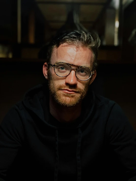 a man with glasses sitting at a table, a character portrait, by Jacob Toorenvliet, unsplash, pewdiepie, low quality photo, standing with a black background, james gleeson