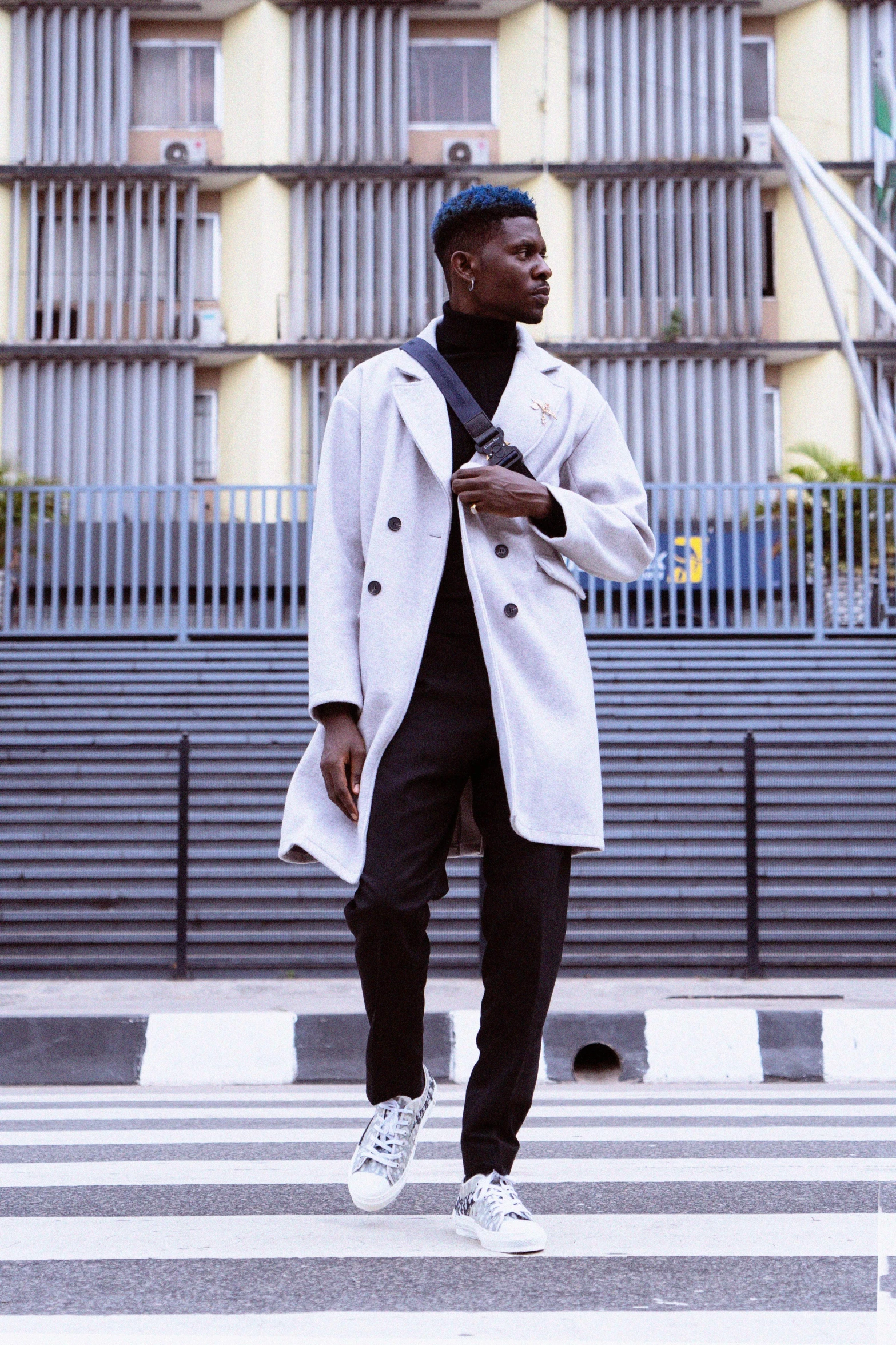 a man walking across a crosswalk in front of a building, an album cover, inspired by Theo Constanté, pexels contest winner, ( ( wearing a long coat ) ), unmistakably kenyan, model posing, satisfied pose