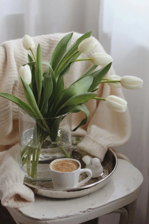 a glass vase filled with white tulips next to a cup of coffee, inspired by Lili Árkayné Sztehló, white sweater, beige, celebration of coffee products, silver，ivory
