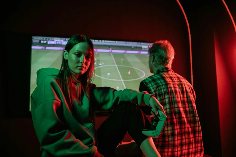 a couple of people sitting in front of a tv, inspired by Elsa Bleda, interactive art, soccer, red and green lighting, woman in streetwear, promotional image