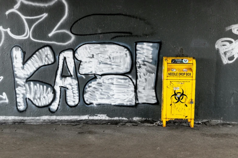 a yellow trash can sitting next to a graffiti covered wall, unsplash, graffiti, meth lab, background image, worksafe, grey
