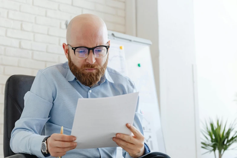 a man sitting in a chair reading a piece of paper, a cartoon, pexels contest winner, ginger bearded man with glasses, people at work, bald, realistic »