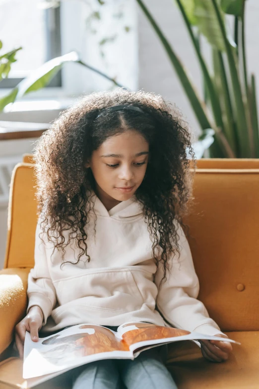 a girl sitting on a couch reading a book, a cartoon, trending on pexels, long afro hair, little kid, wearing a yellow hoodie, grainy