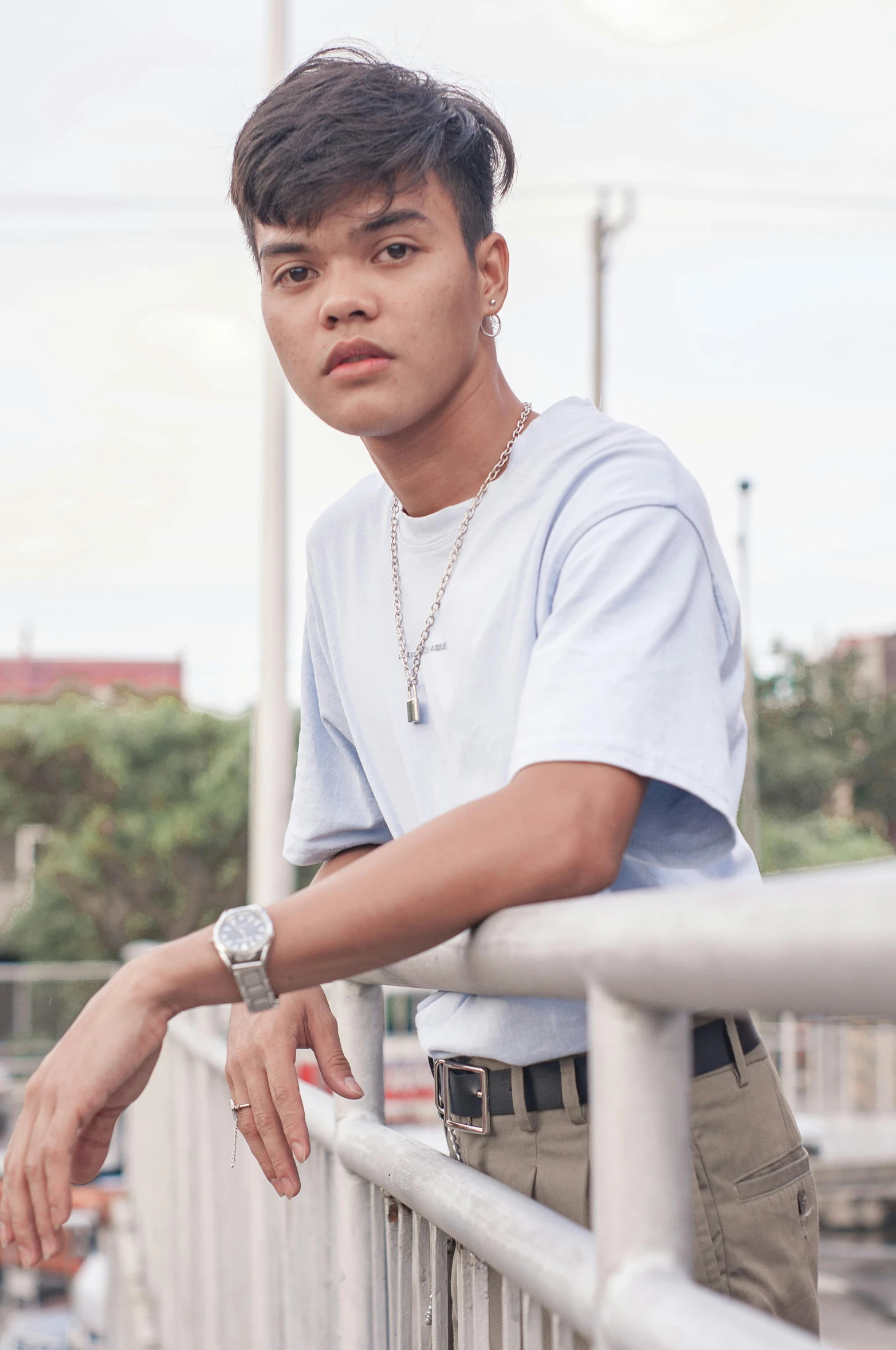 a young man standing on top of a bridge, an album cover, inspired by Jorge Jacinto, trending on pexels, realism, wearing a light blue shirt, jewelry, south east asian with round face, non binary model