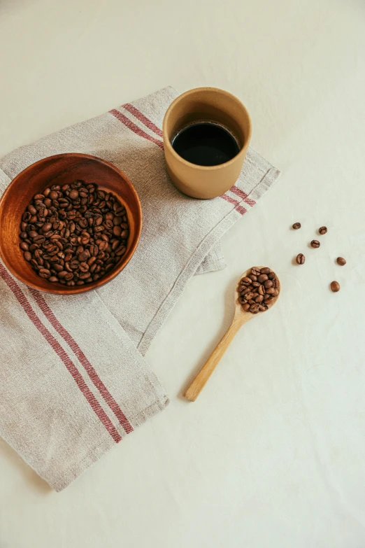 a cup of coffee next to a bowl of coffee beans, a still life, inspired by Ceferí Olivé, trending on unsplash, coffee stain on napkin, spoon placed, detailed product image, detail shot