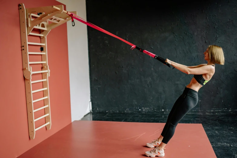 a woman using a pull up bar in a gym, pexels contest winner, red wires wrap around, wooden staff, pink body harness, stretching to walls