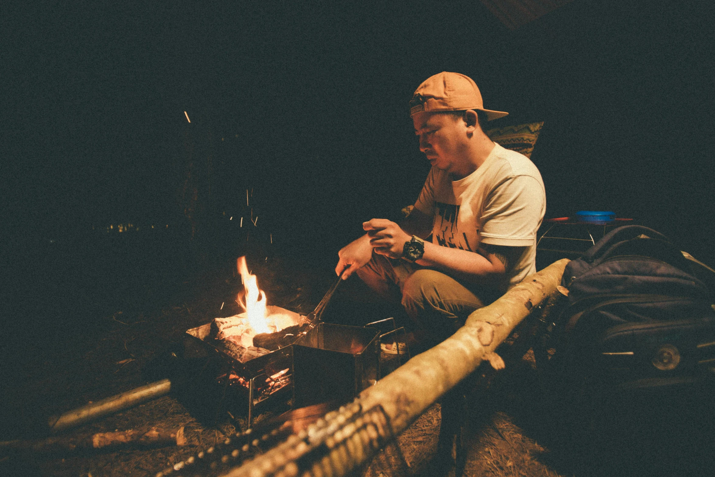 a man sitting on the ground next to a campfire, by Jessie Algie, with some sausages on the fire, darren quach, fishing, nightlife