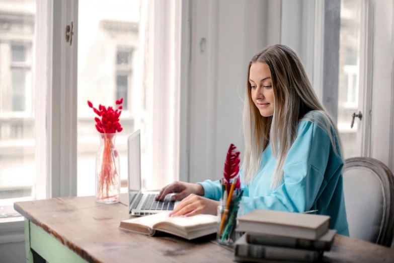 a woman sitting at a table working on a laptop, by Alice Mason, pexels contest winner, a girl with blonde hair, textbooks and books, next to window, thumbnail