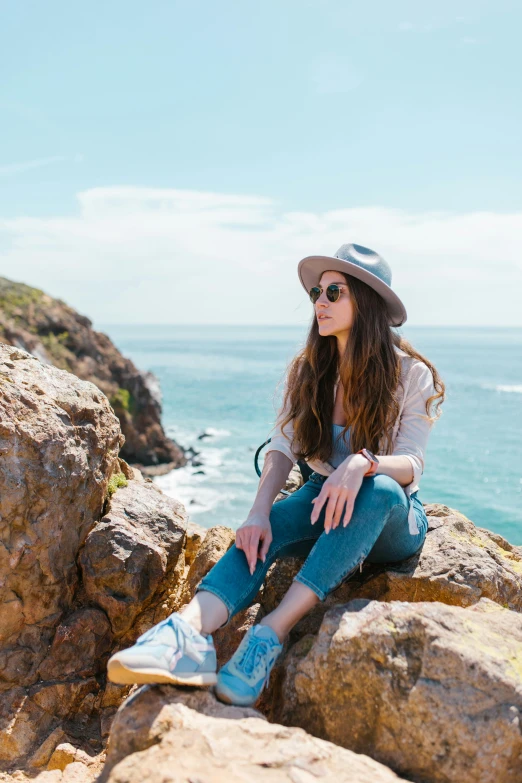 a woman sitting on top of a rock next to the ocean, wearing blue sunglasses, profile image, malibu canyon, wearing white sneakers