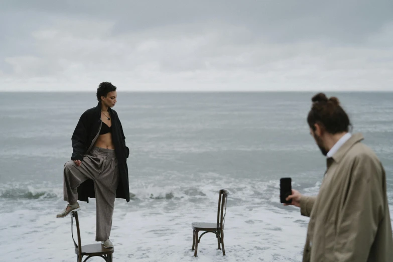 a man sitting on top of a chair next to the ocean, inspired by Peter Lindbergh, unsplash, figuration libre, aurora aksnes and zoë kravitz, standing next to water, meet the actor behind the scenes, goddess checking her phone