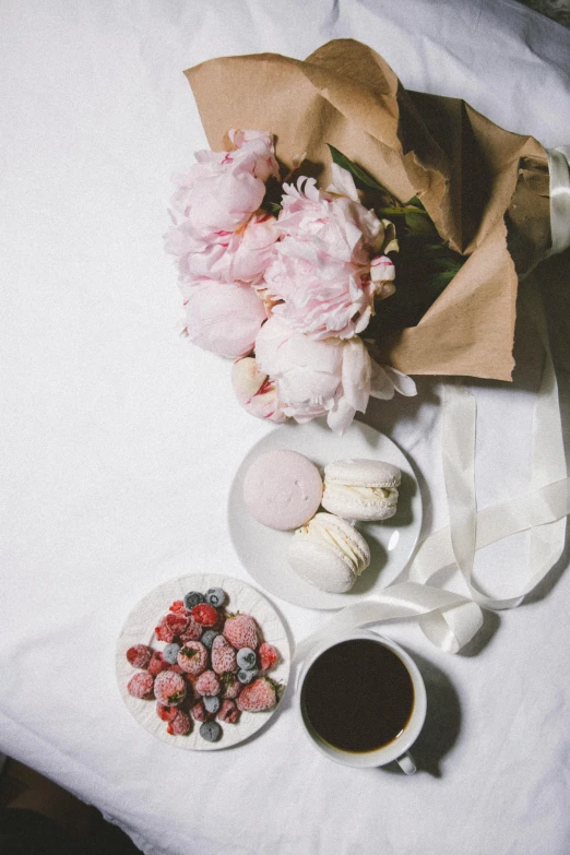 a bouquet of flowers sitting on top of a table next to a cup of coffee, macaron, white and pink cloth, fruit, snacks