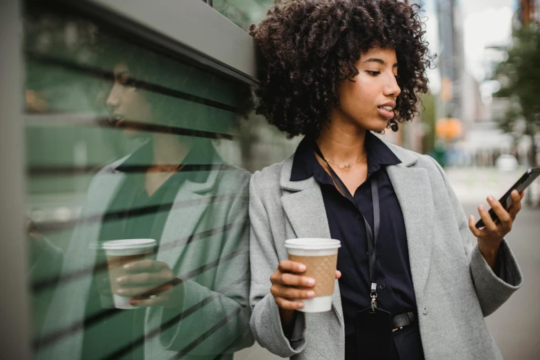 a woman holding a cup of coffee and looking at her cell phone, by Emma Andijewska, trending on pexels, sexy black woman walks past them, wearing jacket and skirt, curly haired, admiring her own reflection