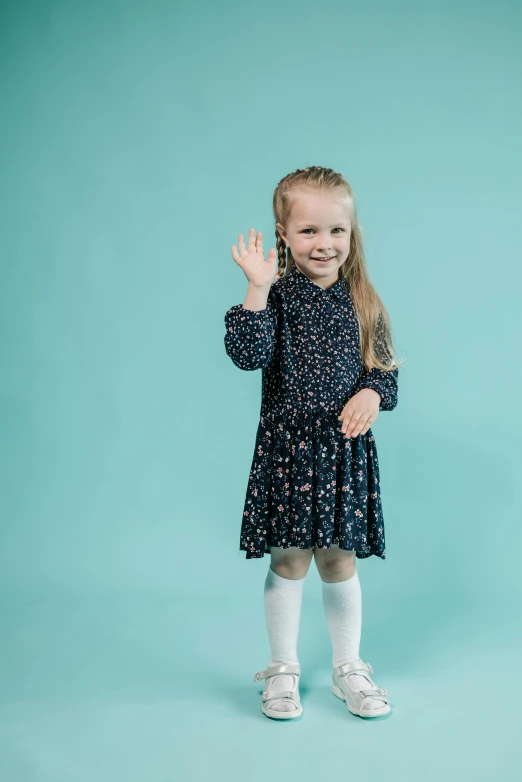 a little girl standing in front of a blue background, pexels, antipodeans, patterned clothing, waving, knobbly knees, school girl in gothic dress