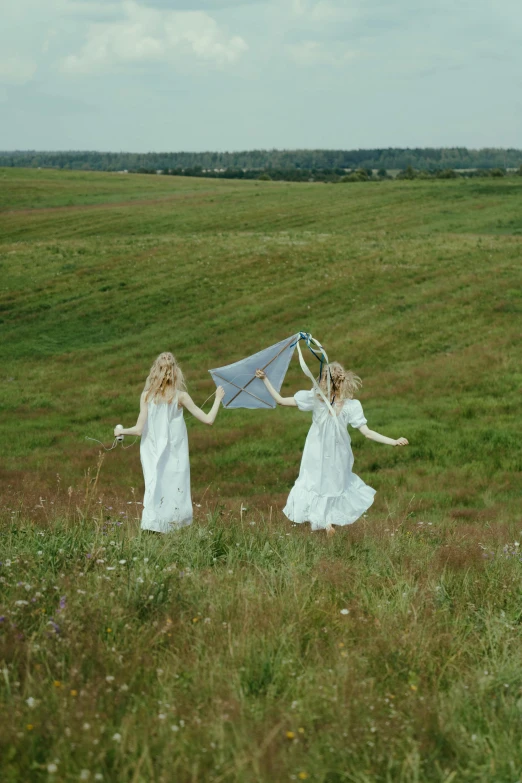 a couple of women standing on top of a lush green field, inspired by Konstantin Somov, unsplash, land art, kites, ignant, white, video still