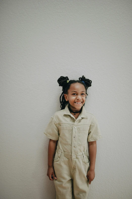 a little girl standing in front of a white wall, by Winona Nelson, pexels contest winner, portrait willow smith, schools, happy kid, overalls