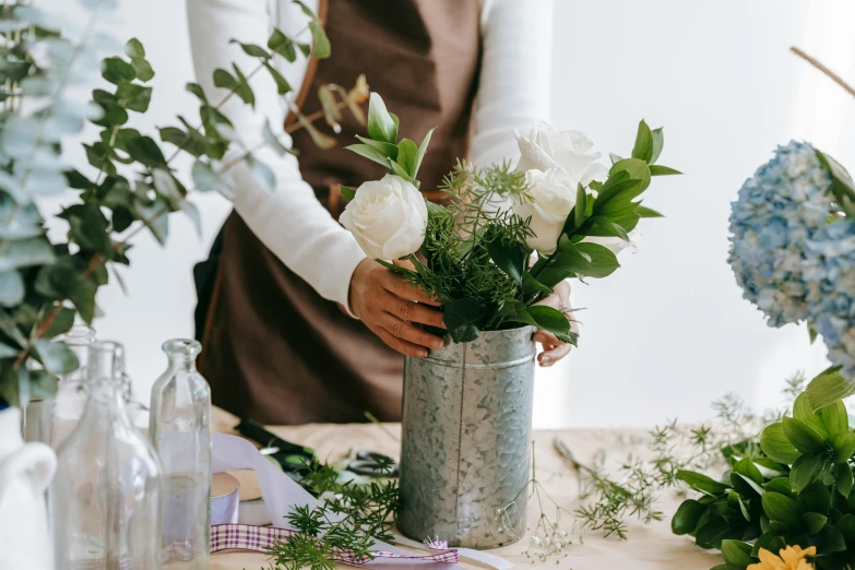 a woman arranging flowers in a vase on a table, trending on pexels, holding a tin can, pale greens and whites, standing elegantly, presenting wares