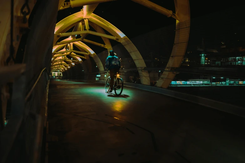 a person riding a bike on a bridge at night, looking towards the camera, an archway, at night time