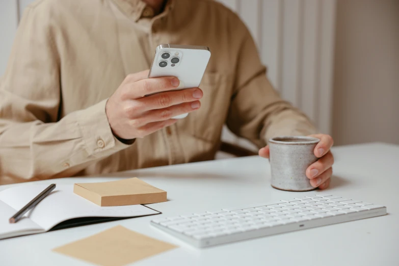 a man sitting at a desk using a cell phone, trending on pexels, realism, with a white mug, brown and white color scheme, holding a stuff, bulky build