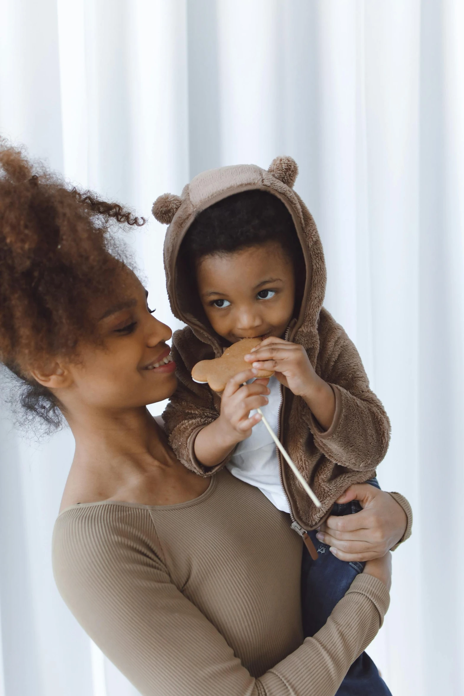 a woman holding a child in her arms, by Lily Delissa Joseph, pexels contest winner, afro comb, beige hoodie, having a snack, slightly minimal