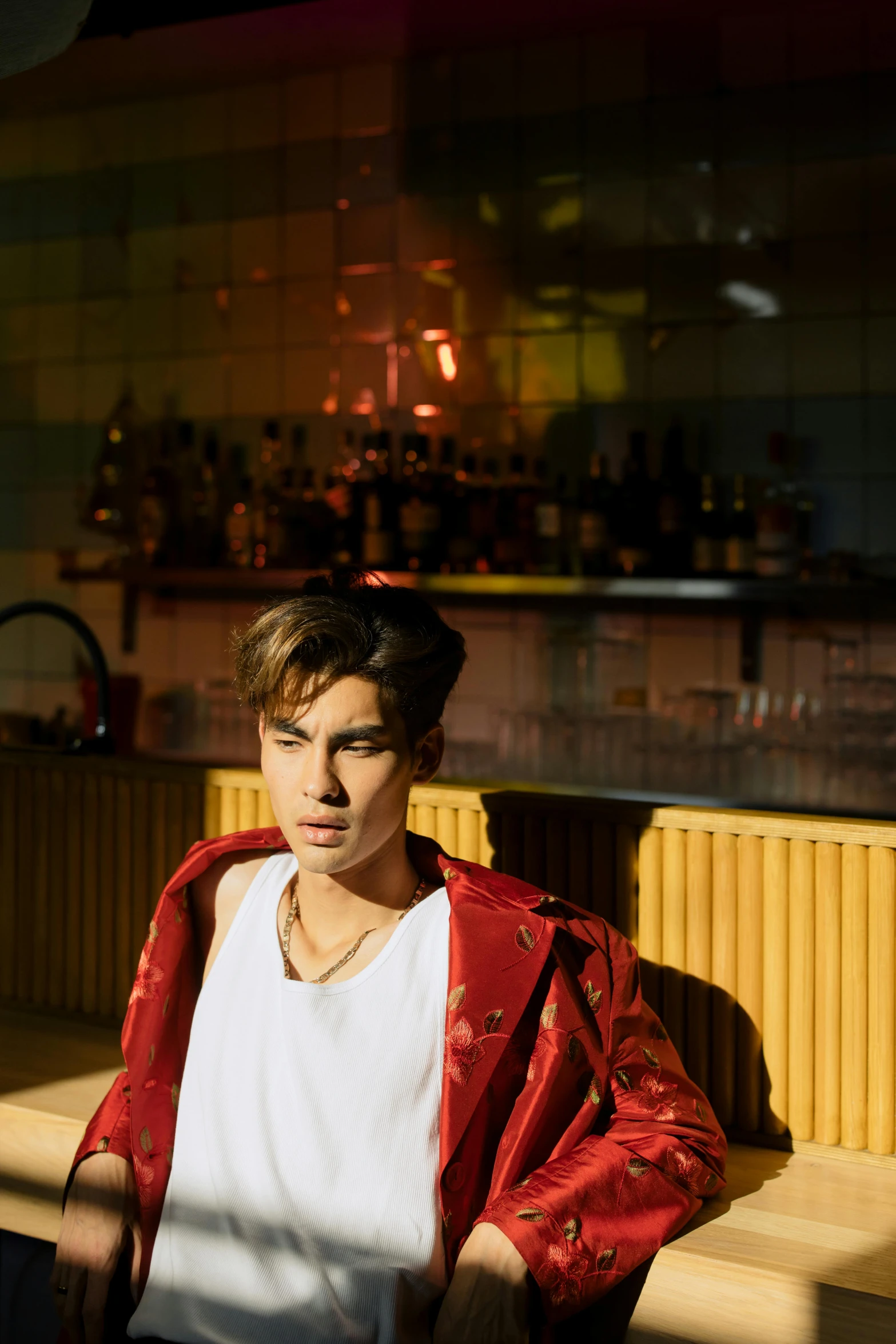 a man sitting on a bench in front of a bar, an album cover, unsplash, androgynous male, portrait of archie andrews, standing in a restaurant, red cloth around his shoulders