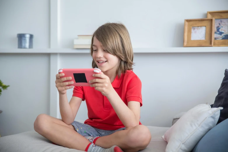 a little girl sitting on top of a bed holding a cell phone, holding a nintendo switch, red shirt, coral red, uk