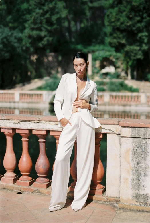 a woman in a white suit sitting on a railing, inspired by Luis Paret y Alcazar, unsplash, baroque, tall and slender, tan suit, lush surroundings, at a fashion shoot