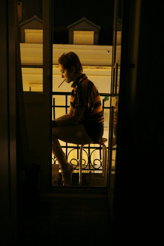 a person sitting on a balcony at night, with a cigarette in its mouth, leaving a room, afternoon, facing away