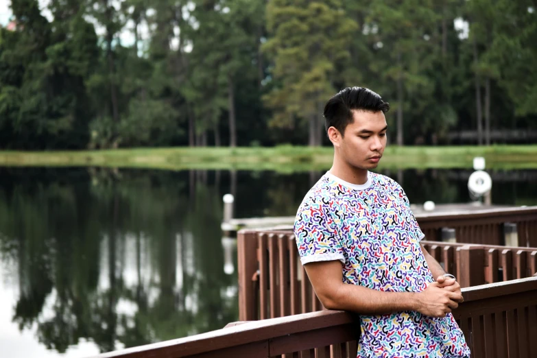 a man standing on a bridge next to a body of water, a portrait, inspired by Reuben Tam, unsplash, visual art, multicolored tshirt art, in front of a forest background, patterned clothing, florida man