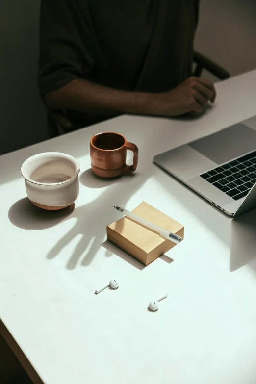 a laptop computer sitting on top of a white table, by Pablo Rey, cigarrette boxes at the table, holding pencil, with a white mug, clay material