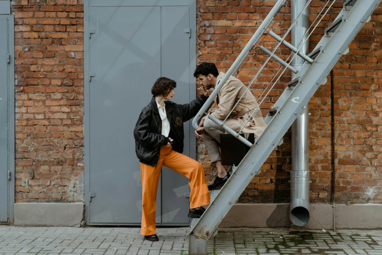 a man and a woman climbing up a set of stairs, a photo, trending on pexels, black and orange coat, flirting, wearing pants, industrial colours