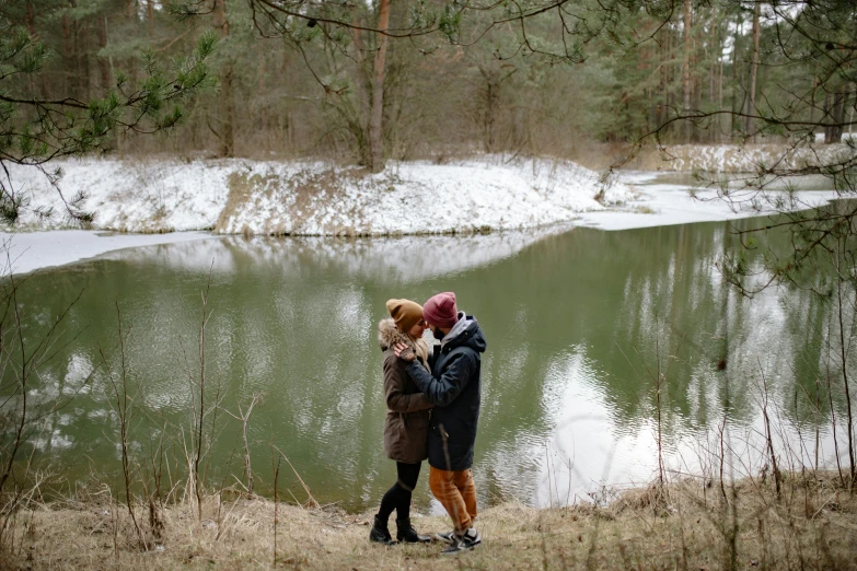 a couple of people standing next to a body of water, by Grytė Pintukaitė, pexels contest winner, spring winter nature melted snow, lesbian embrace, forest with lake, really realistic