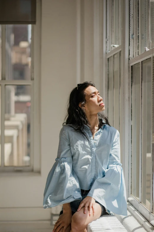 a woman sitting on a window sill looking out a window, an album cover, inspired by Fei Danxu, pexels contest winner, wearing a light blue shirt, voluminous sleeves, bao pham, androgynous person