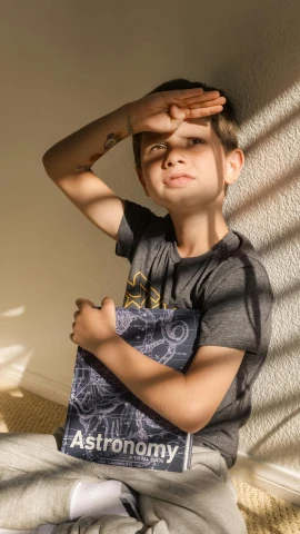 a young boy sitting on a bed holding a book, by Pamela Drew, pexels contest winner, graffiti, wearing a muscle tee shirt, solar punk product photo, holding a gold bag, some sun light ray