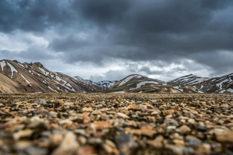 a rocky field with mountains in the background, by Jesper Knudsen, unsplash contest winner, gravel and scree ground, storm clouds in the distance, earth colors, desolate arctic landscape