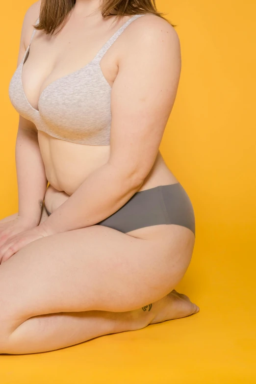 a woman sitting on the ground posing for a picture, a colorized photo, by Austin English, trending on unsplash, figuration libre, she has a jiggly fat round belly, underwear ad, plain background, yellow