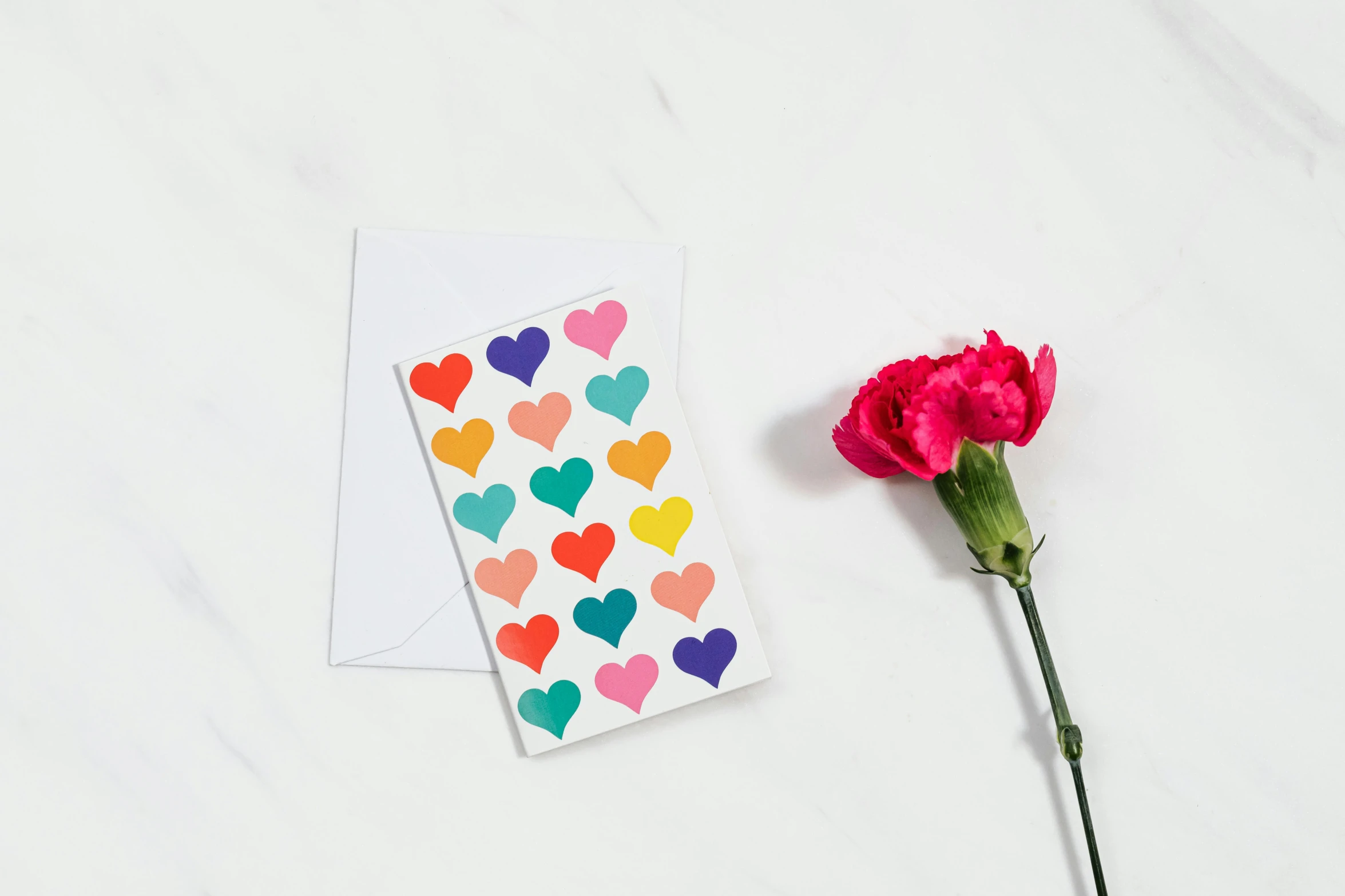 a card sitting on top of a table next to a flower, many hearts, product shot, multicoloured, plain background