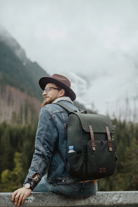 a man with a backpack sitting on a fence, by Jessie Algie, pexels contest winner, square backpack, wearing a travel hat, forrest in the background, on a gray background